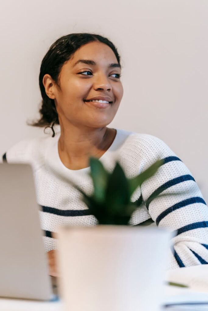 Happy ethnic woman in casual outfit working remotely with netbook at table near potted green plant in room and looking away